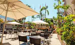 Bilde av en uteplass med bord, stoler og parasoller i et tropisk miljø. (AI-generert tekst)