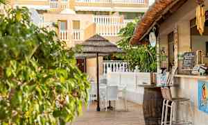 Utendørs serveringsområde med stoler og en parasoll i nærheten av en bygning med gul fasade og hvite rekkverk. (AI-generert tekst)