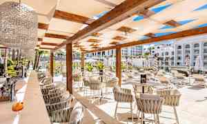 En utebar med stoler og bord under en pergola, omgitt av solsenger og grøntområder. (AI-generert tekst)