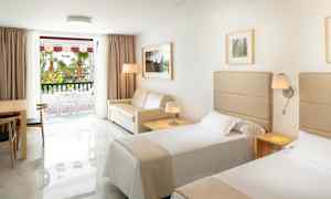 Hotellrom med to enkeltsenger, en sofa, et bord med stoler og et vindu mot en balkong. (AI-generert tekst)