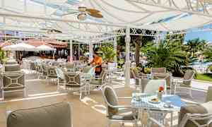 Utendørs restaurant med solskjerming, bord og stoler, samt grøntområder i bakgrunnen. (AI-generert tekst)