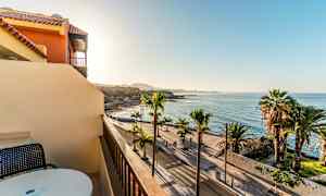 Utsikt fra en balkong mot en palmekantet strandpromenade og havet under en klar himmel. (AI-generert tekst)