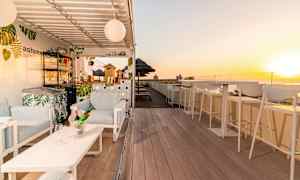 En solfylt takterrasse med stoler, bar og bord, samt solnedgang i bakgrunnen. (AI-generert tekst)