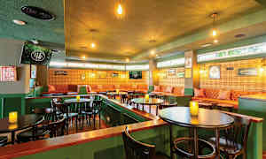 En koselig innredet kafé eller pub med grønne vegger, belyst av hengende lamper, brune bord og stoler, samt en sittegruppe med stoppede sofaer og veggdekorasjoner. (AI-generert tekst)