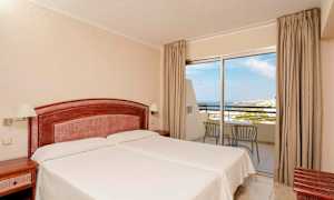 Et hotellrom med to enkeltsenger, en trehodegavl og nattbord. Et glassparti med utsikt mot havet og balkongmøbler. (AI-generert tekst)