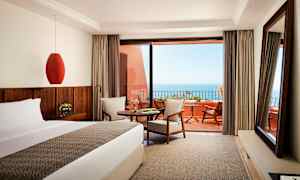 Et hotellrom med seng, bord og sitteområde, med utsikt mot havet fra en terrasse. (AI-generert tekst)