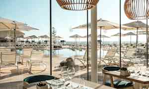 Utsikt fra en restaurant med bord og glasservise mot en terrasse med solsenger og parasoller ved et basseng. (AI-generert tekst)