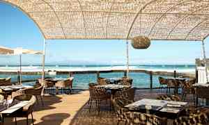 En utendørsrestaurant ved sjøen med rottingsstoler, trebord og en halvtransparent takstruktur. (AI-generert tekst)