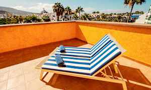 Utsikt fra en solterrasse med to solsenger med stripete blå og hvite puter samt håndklær på dem, omgitt av palmer og bygninger. (AI-generert tekst)