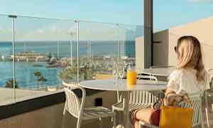 En person sitter ved et bord med et glass og en karaffel appelsinjuice med utsikt over en småbåthavn. (AI-generert tekst)