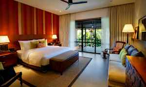 Et romslig soverom med en stor seng, to nattbord med lamper, en sofa, en lenestol og en balkong med utsikt over frodig vegetasjon. (AI-generert tekst)