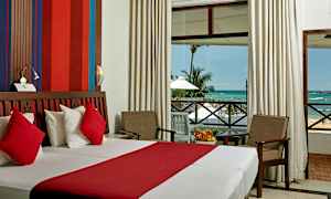 Et hotellrom med en dobbeltseng med rødt og hvitt sengetøy, en sittegruppe med to stoler og et bord, og en stor verandadør som viser utsikten mot havet og palmetrær. (AI-generert tekst)