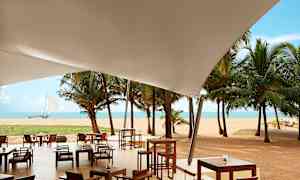 Utendørs serveringsområde under et hvitt tak med utsikt mot sandstrand, palmer og hav. (AI-generert tekst)