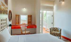 Et lyst hotellrom med en hvit seng, et rødt bord med en skrivebordsstol og skodder ved vinduet. En mønstret lenestol står på den andre siden av rommet. (AI-generert tekst)