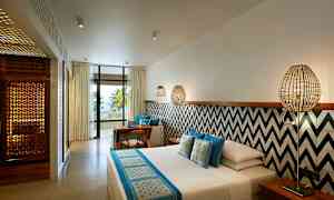 Et hotellrom med moderne innredning, seng med blå og hvite tekstiler, vegg med geometriske mønstre, sittegruppe og en balkong med utsikt mot palmer og havet. (AI-generert tekst)