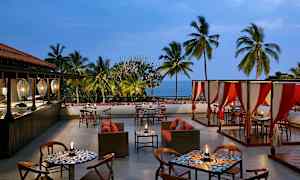En utendørsrestaurant med bord, stoler og sofaer, omgitt av palmer med havet synlig i bakgrunnen. (AI-generert tekst)