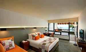 Hotellrom med moderne innredning, med en dobbeltseng med fargerike puter og sengeteppe. Et skrivebord, en sofa og en TV kompletterer rommet. Det finnes en balkong med utsikt over havet. (AI-generert tekst)