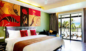 Hotellrommet med en stor seng, fargerike puter, en sittegruppe og en balkong med utsikt over en sandstrand omgitt av palmer. (AI-generert tekst)