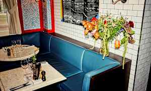 En kafe eller restaurant med et bord og blå sitteplasser. Bordet er dekket med vinglass og krydderkverner. En blomsterbukett står ved siden av på et sidebord. (AI-generert tekst)