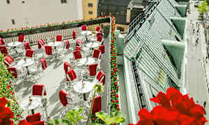 En takterrasse med hvite bord og stoler, dekorert med røde tepper. Rundt er det blomsterkasser med røde blomster og utsikt over tak og gater. (AI-generert tekst)