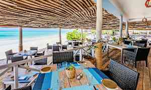 Uteservering med tak av flettet bast ved en sandstrand og blått hav. (AI-generert tekst)