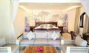 Et hotellrom med sofa og lenestoler foran en stor seng med pynteputer og tropisk bakgrunn. (AI-generert tekst)