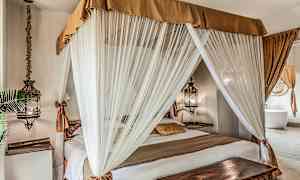 Soverom med en himmelseng omgitt av hvite gardiner og dekorert med varme tonede tekstiler. (AI-generert tekst)