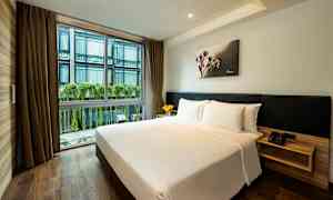 Et hotellrom med en dobbeltseng, lyst sengetøy og tregulv. Store vinduer med utsikt mot en bygning med planter hengende langs fasaden. (AI-generert tekst)