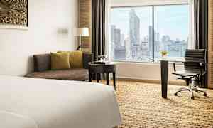 Bilde av et hotellrom med en seng, en sofa, et skrivebord og en stol. Et stort vindu viser en utsikt over en by. (AI-generert tekst)