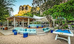 En utendørs terrasse med sittegrupper og bord under solbeskyttelsestak, omgitt av grøntområder og med en klippe i bakgrunnen. (AI-generert tekst)