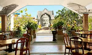 Utendørs servering med trestoler og bord, omgitt av grøntområder og dekorative planter. En arkitektonisk struktur er synlig i bakgrunnen. (AI-generert tekst)