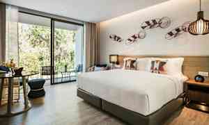 Bilde av et moderne hotellrom med en stor seng, dekorative puter, sittemøbler og en glassvegg som vender mot en terrasse og grøntområder utenfor. (AI-generert tekst)