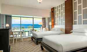 Et hotellrom med to separate enkeltsenger, moderne møbler og et lyst design. Et stort vindu med utsikt over havet. (AI-generert tekst)