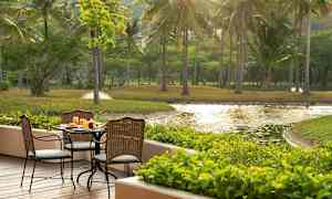 Uteservering med bord og stoler på en treterrasse, omgitt av grøntområder og utsikt over en dam. (AI-generert tekst)