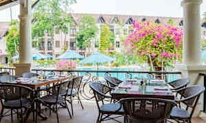 Utendørs servering med bord og stoler ved et basseng. I bakgrunnen vises bygninger, parasoller og grøntområder. (AI-generert tekst)