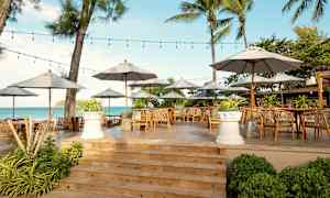 En utendørsrestaurant med bord, stoler og parasoller på en treplattform nær sjøen. (AI-generert tekst)