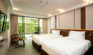 Et hotellrom med to enkeltsenger med hvitt sengetøy, et vindu med utsikt over en hage og et trebord med en stol ved siden av. (AI-generert tekst)