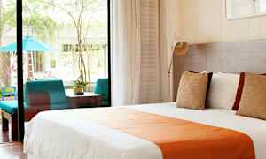 Bilde av et lyst hotellrom med en stor seng dekket med hvite og oransje tekstiler, store vinduer som vender ut mot en terrasse med sittegruppe og planter. (AI-generert tekst)