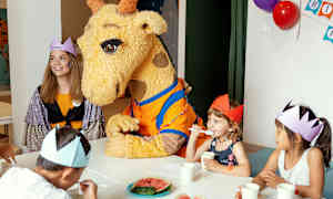 En giraffmaskot sitter ved et bord med barn som har på seg papirkrone. (AI-generert tekst)