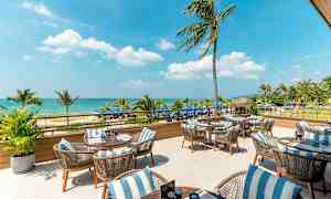 Utendørs servering med bord og stoler, hav og palmer i bakgrunnen. (AI-generert tekst)