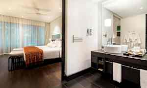 Et hotellrom med sovedel og bad. Sovedelen har en seng med hvitt sengetøy og oransje teppe. Badet har servant med speil. (AI-generert tekst)