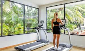 En person ser på skjermen til en tredemølle i et treningsstudio med store vinduer og utsikt mot grøntområder. (AI-generert tekst)
