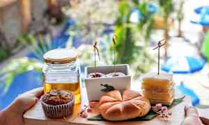 En brett med forskjellige bakverk og en glasskrukke med honning, holdt foran en uskarp bakgrunn med planter og parasoller. (AI-generert tekst)