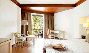 Et hotellrom med hvitt skrivebord, seng og sittemøbler, samt balkong med utsikt over trær. (AI-generert tekst)