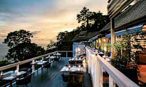 Uteservering med bord og stoler om kvelden, med utsikt over trær og hav. (AI-generert tekst)