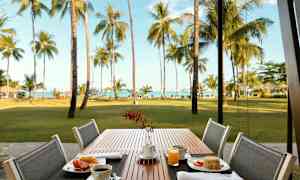 Uteservering med en frokost ved et bord. Bakgrunnen viser en plen, palmer og hav. (AI-generert tekst)