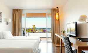 Et hotellrom med to senger, skrivebord og utsikt mot en balkong med hav i bakgrunnen. (AI-generert tekst)