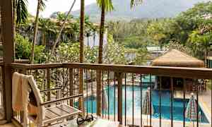 Utsikt fra en balkong over et basseng omgitt av palmer og grønn vegetasjon, med en stol og et bord i forgrunnen. (AI-generert tekst)