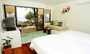 Et lyst hotellrom med seng, sofa og en stor glassdør som leder ut til en terrasse med hengekøye og utsikt over havet. (AI-generert tekst)