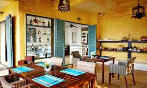 Et rom med gule vegger og turkise dører, innredet med trebord og rottingstoler. Hyller med flasker og serveringsartikler er synlige. (AI-generert tekst)